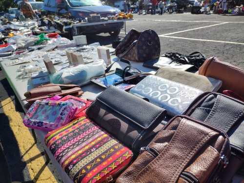 A variety of handbags and wallets displayed on a table at a flea market, with cars and other items in the background.