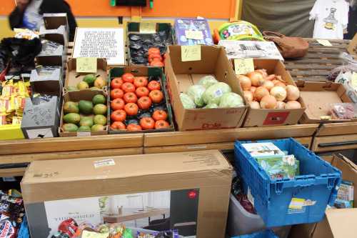 A market stall displaying various fruits and vegetables in cardboard boxes, with prices visible on signs.