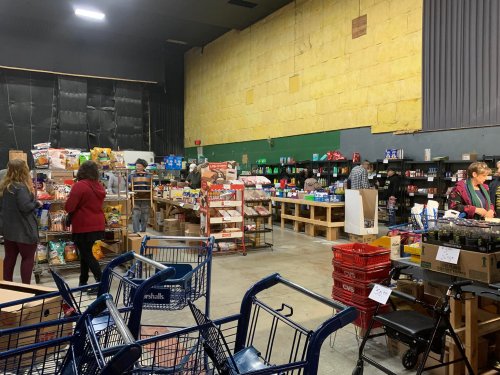 A busy community food pantry with shelves of groceries and people shopping, carts in the foreground.
