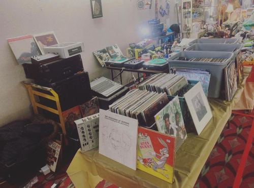 A collection of vinyl records displayed on tables, with various music equipment and items in a market setting.