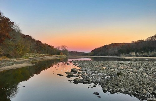 A serene river at sunset, reflecting colorful skies and surrounded by trees and rocky shores.