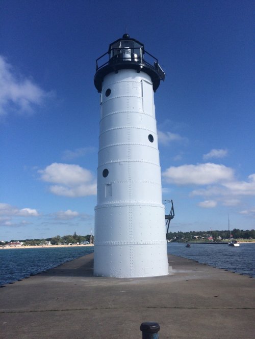 A tall white lighthouse stands on a pier, surrounded by water and blue skies with a few clouds.