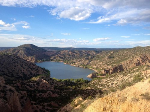 A scenic view of a lake surrounded by hills and mountains under a partly cloudy sky.