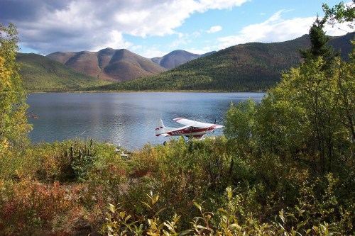 A seaplane rests by a tranquil lake surrounded by mountains and lush greenery under a partly cloudy sky.