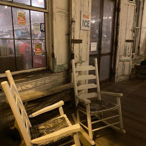 Two rustic wooden chairs sit on a porch beside a window, with a sign for "Bag Ice" visible nearby.