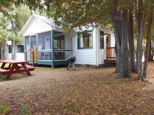 A cozy white cottage surrounded by trees, featuring a screened porch, picnic table, and a small grill in the yard.