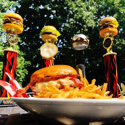 A delicious burger with fries in front of towering trophy burgers, set against a green background.