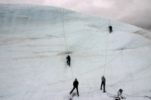Four climbers ascend a large glacier, using ropes for support against a backdrop of cloudy skies.