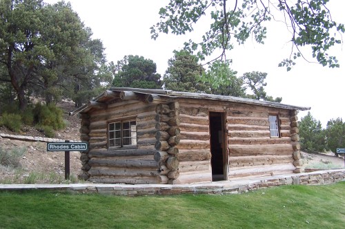 A rustic log cabin with a sign reading "Rhodes Cabin," surrounded by trees and grassy terrain.