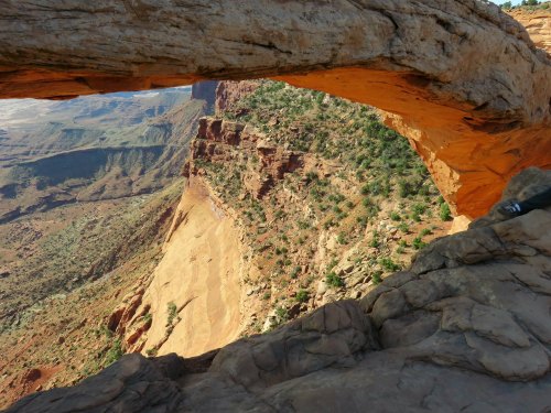 A natural stone arch overlooks a rugged canyon landscape with green vegetation and rocky cliffs in the background.