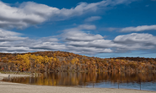 A serene lake surrounded by autumn foliage and a cloudy blue sky, reflecting vibrant colors on the water's surface.