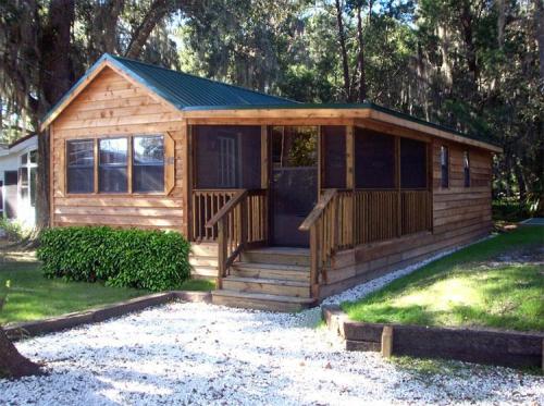 A wooden cabin with a green roof, front porch, and surrounded by greenery and gravel pathway.
