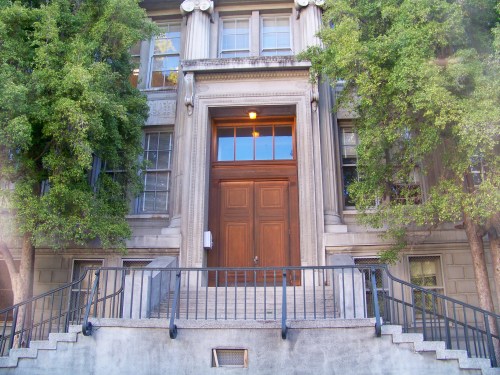 Front view of a historic building with a large wooden door, flanked by trees and a staircase leading up to it.