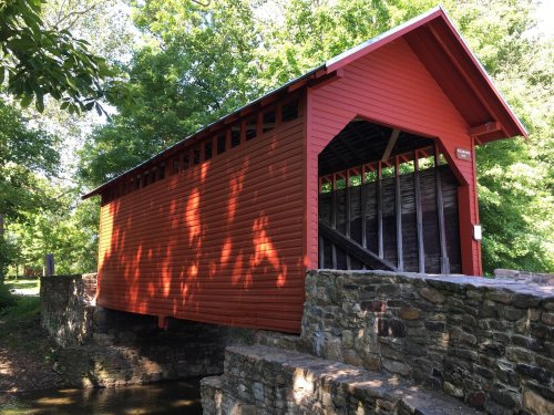 A red covered bridge with a stone foundation, surrounded by lush green trees and dappled sunlight.