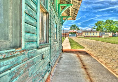 A weathered green building with a sidewalk leading to a clear sky and distant structures in a rural setting.