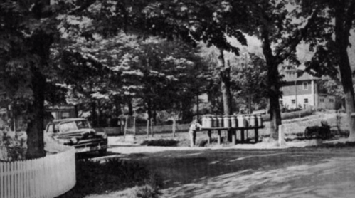 A black-and-white photo of a street scene with trees, a parked car, and a person near a table with barrels.