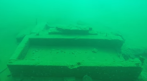 Underwater view of a submerged structure covered in sediment, with a greenish tint from the water.