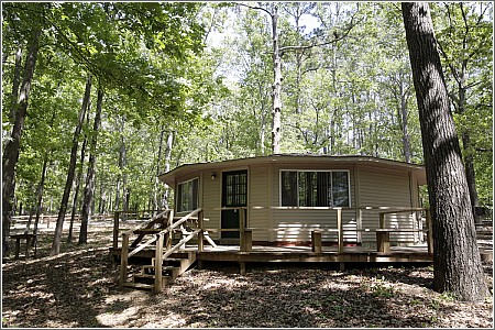 A round cabin surrounded by trees, featuring a wooden deck and large windows in a serene forest setting.