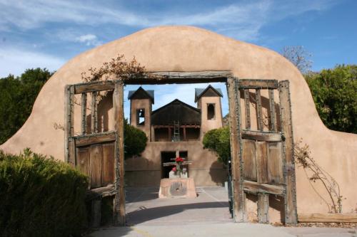 A rustic adobe archway with wooden doors leads to a courtyard featuring greenery and a building with towers.