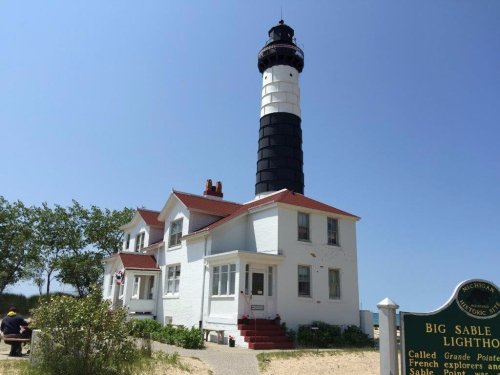 A tall black and white lighthouse beside a white building, surrounded by greenery and a clear blue sky.