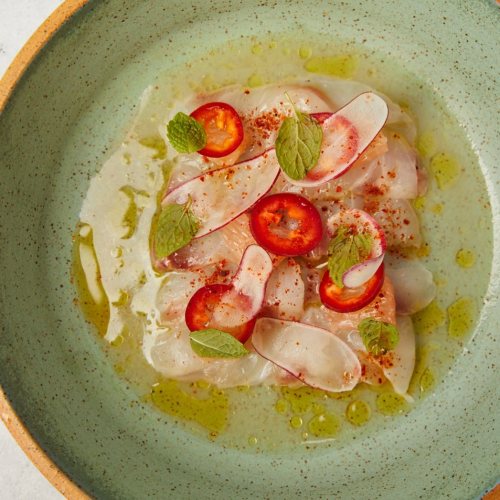 A green bowl filled with thinly sliced fish, red chili slices, herbs, and a drizzle of olive oil.