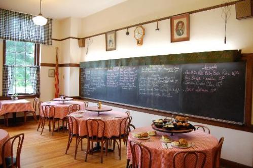 A cozy dining room with round tables, checkered tablecloths, and a chalkboard menu on the wall.