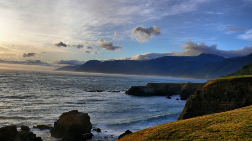 A scenic coastal view with rocky cliffs, rolling waves, and mountains under a cloudy sky at sunset.
