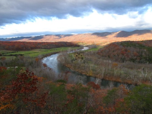 A scenic view of a winding river surrounded by autumn foliage and rolling hills under a cloudy sky.