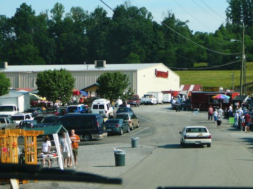 A busy outdoor market scene with vendors, cars, and people gathered near a large building and green trees in the background.