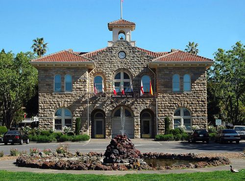 Historic stone city hall building with a clock tower, surrounded by greenery and a small fountain in the foreground.