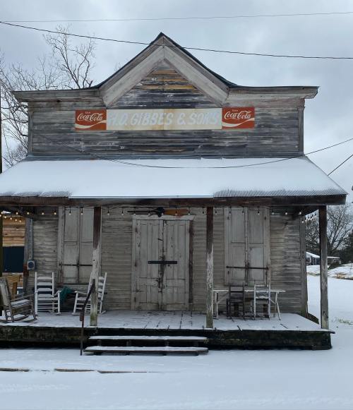 Old wooden building with "T.D. Gibbs & Sons" sign, covered in snow, featuring rocking chairs on the porch.