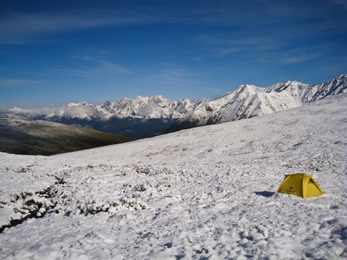 A yellow tent sits on a snowy landscape, surrounded by majestic mountains under a clear blue sky.