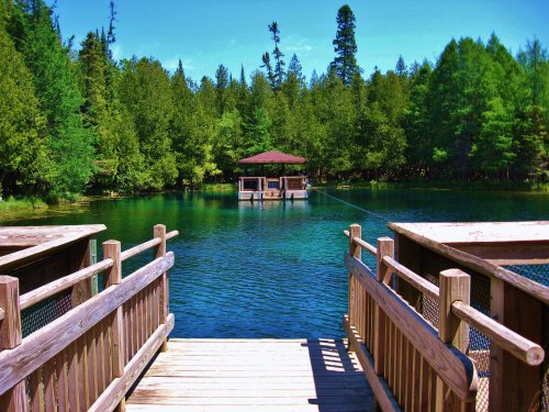A wooden dock leads to a serene lake surrounded by lush green trees and a small gazebo on the water.