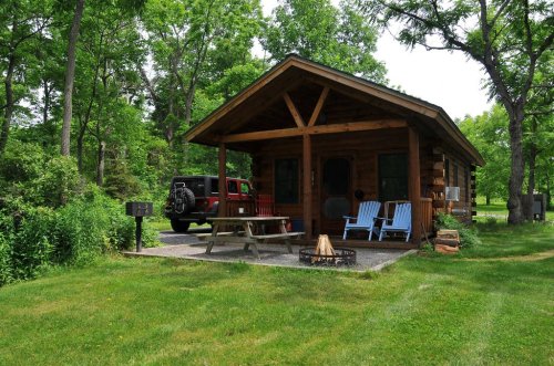 A cozy log cabin surrounded by greenery, featuring a picnic table, fire pit, and two blue chairs on the porch.