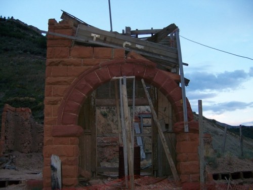 Ruined stone structure with a collapsed roof, surrounded by wooden supports and debris against a twilight sky.