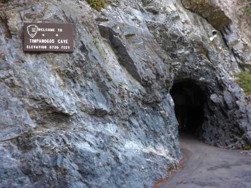 Entrance to Timpanogos Cave, featuring a sign and rocky surroundings, with a dark tunnel visible inside.