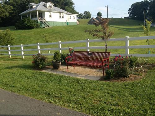 A red bench surrounded by flowers and plants, with a white fence and a house on a grassy hill in the background.