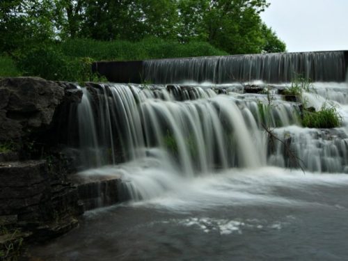 A serene waterfall cascading over rocks, surrounded by lush greenery and a calm body of water.