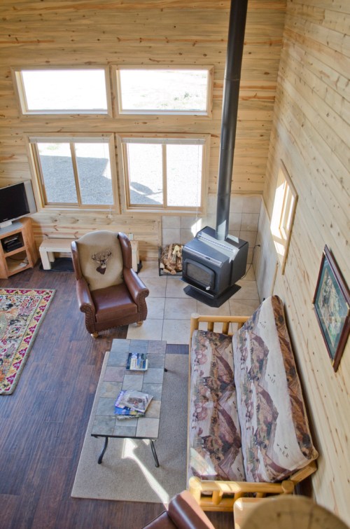 Cozy wooden cabin interior with a sofa, armchair, coffee table, and a wood stove, featuring large windows for natural light.