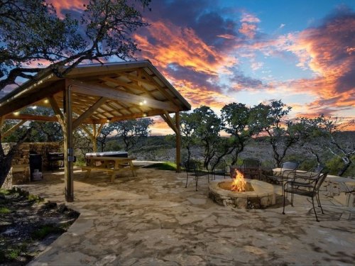 A cozy outdoor space with a fire pit, picnic table, and stunning sunset sky surrounded by trees.