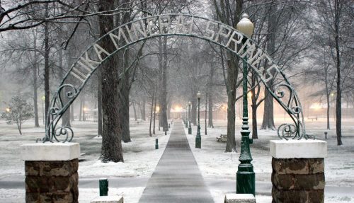 Snow-covered pathway leading through an archway in a park, surrounded by trees and soft lighting.