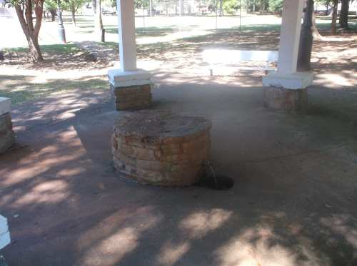 A circular stone well with a small opening, surrounded by a shaded park area with trees in the background.