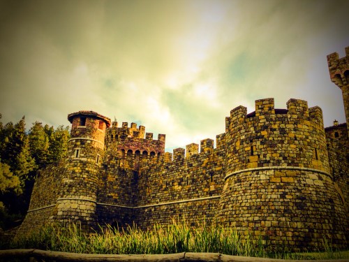 A stone castle with tall towers and a cloudy sky in the background, surrounded by greenery.