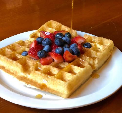 A golden waffle topped with fresh strawberries and blueberries, drizzled with syrup, on a white plate.