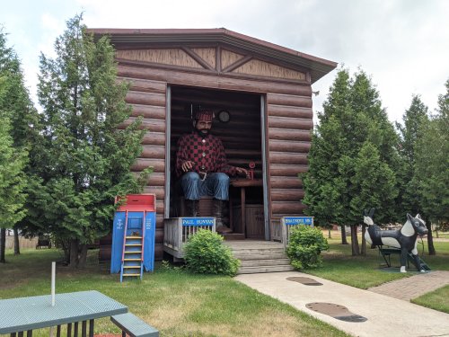 A large statue of Paul Bunyan sits in a log cabin, surrounded by trees and outdoor furniture.