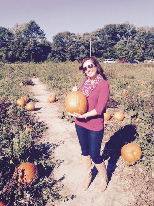 A woman in a maroon top and sunglasses holds a pumpkin in a field filled with pumpkins and greenery.