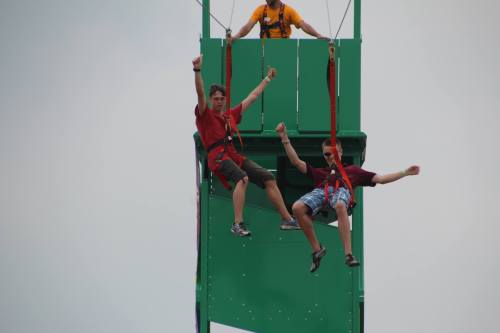 Two people in harnesses are suspended from a tall green structure, smiling and raising their arms in excitement.