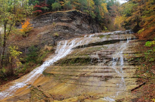 A cascading waterfall flows down layered rock formations, surrounded by autumn foliage in a serene forest setting.