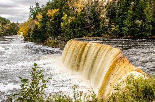 A waterfall cascades over a rocky ledge, surrounded by autumn foliage and a flowing river.