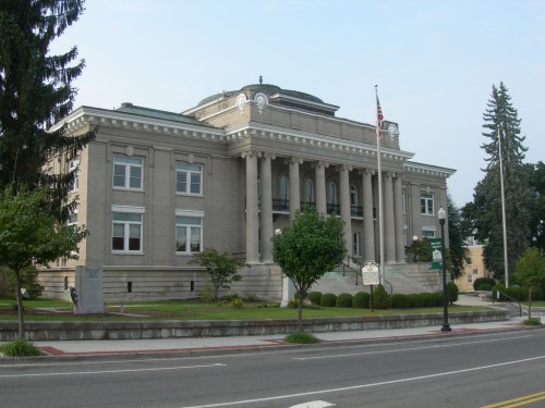 Historic building with columns, flag, and landscaped greenery, set against a clear sky.